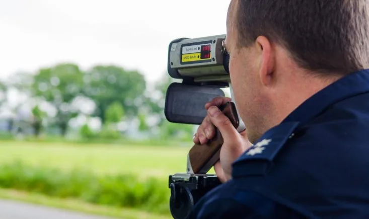 Polizeibeamter visiert mit einem Laserhandmessgerät auf einem Stativ ein Fahrzeug an, im Display stehen Entfernung und Geschwindigkeit