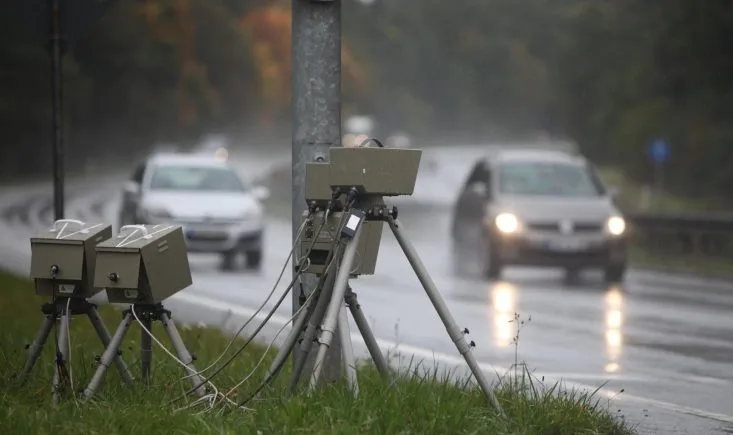 Zwei Messeinheiten auf Stativen stehen bei Regen am Rand einer Landstraße, im Hintergrund fahren zwei Autos mit eingeschaltetem Licht
